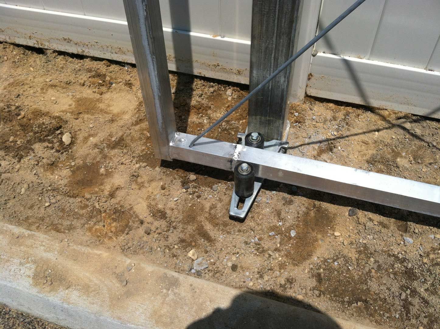 Metal gate frame attached to a post with bracing wires, set in dirt and near a white fence.