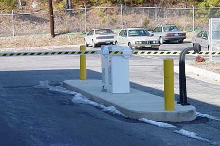 Parking lot gate with striped arm, two yellow posts, and several cars in the background.