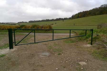 A green metal gate blocks a dirt road in front of a green hillside under a cloudy sky.