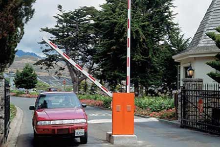 Red car at open security gate, entering a gated community. Orange barrier arm with red and white stripes.