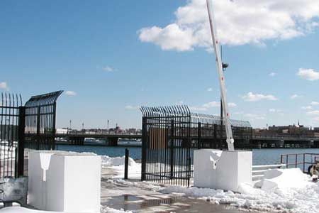 Black fence and gate with razor wire, white blocks, barrier arm, snow, and bridge over water under a blue sky.