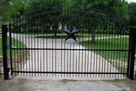 Black metal gate with Texas star, arched top, driveway, and green grass.