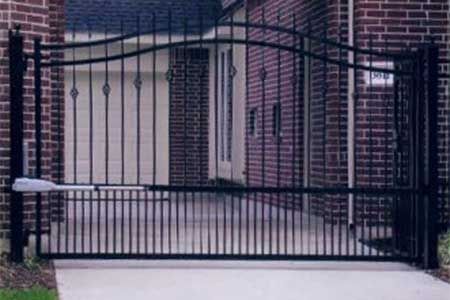 Black metal driveway gate with vertical bars and curved top, opening to a brick home.