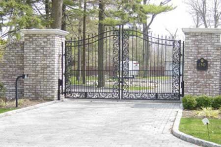 Ornate black iron gates between stone pillars, leading to a driveway, trees in the background.
