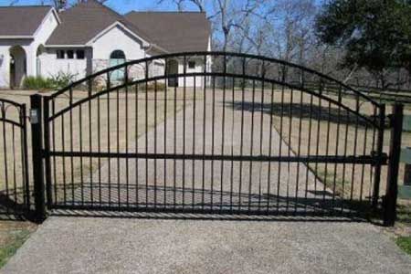 Black wrought-iron driveway gate in front of a house, arched top, closed on a concrete driveway.