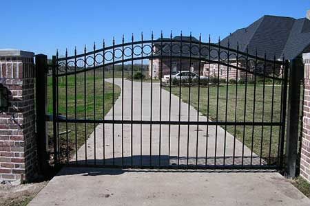 Black wrought-iron gate in front of a concrete driveway leading to a house. Brick pillars frame the gate.