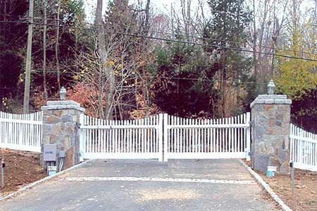 White gated driveway entrance with stone pillars, picket fence, and wooded background.