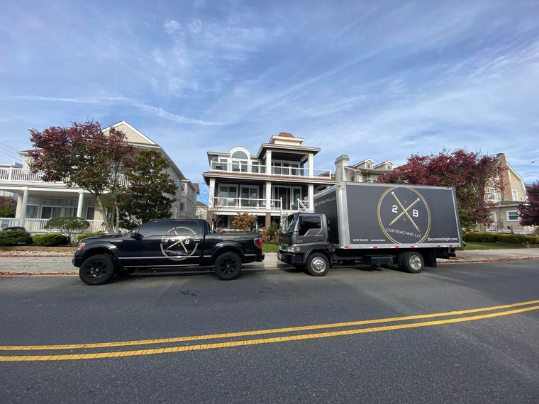 Black truck and box truck parked on a street in front of a house. Cloudy blue sky above.