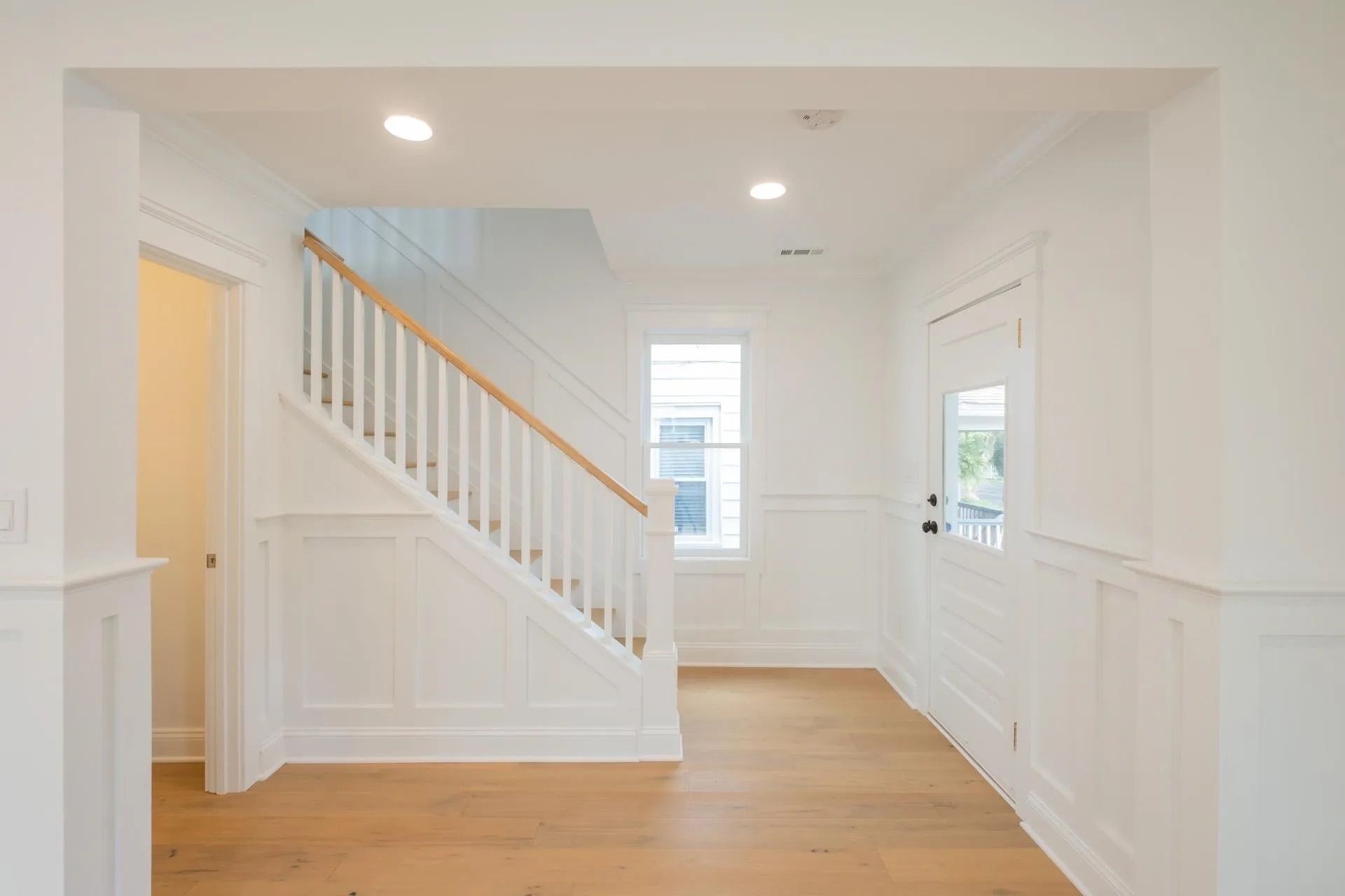 White interior hallway with staircase, hardwood floors, and a window.