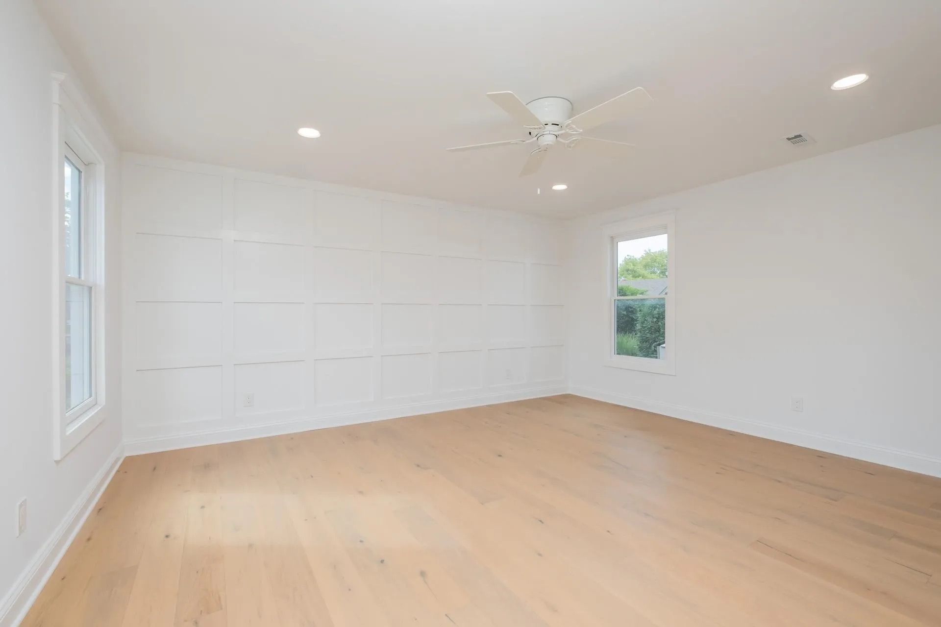 Empty, bright white room with light wood floors, two windows, and a ceiling fan.