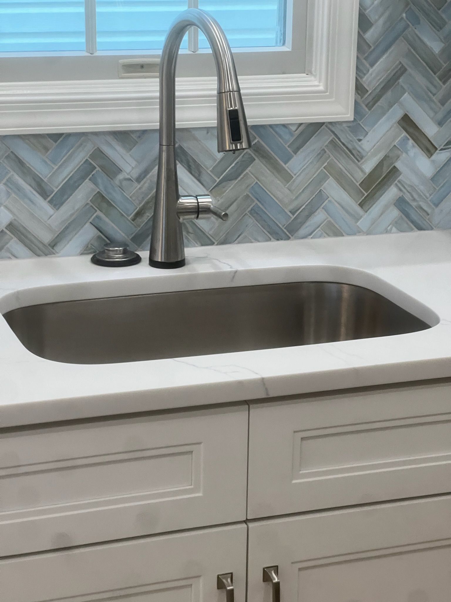 Stainless steel kitchen sink with faucet, white countertop, and herringbone backsplash.