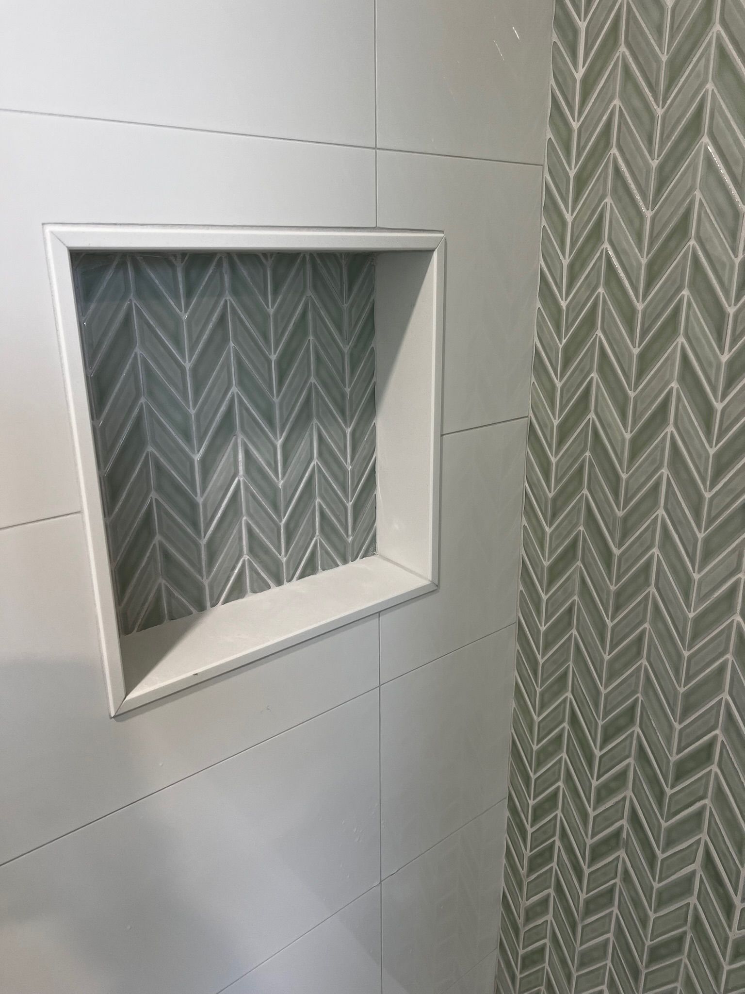 Shower wall with built-in shelf. White walls with a gray herringbone-patterned tile niche and accent wall.