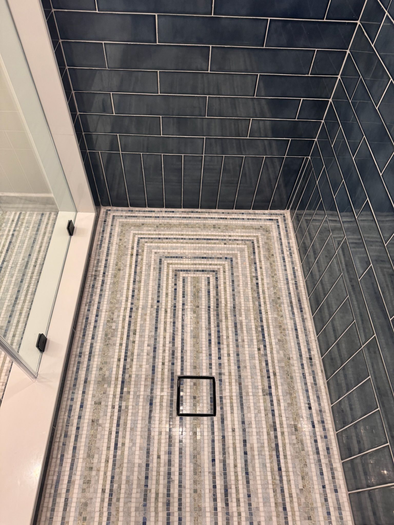 Shower floor with patterned mosaic tiles leading to a square drain, with dark blue rectangular wall tiles.