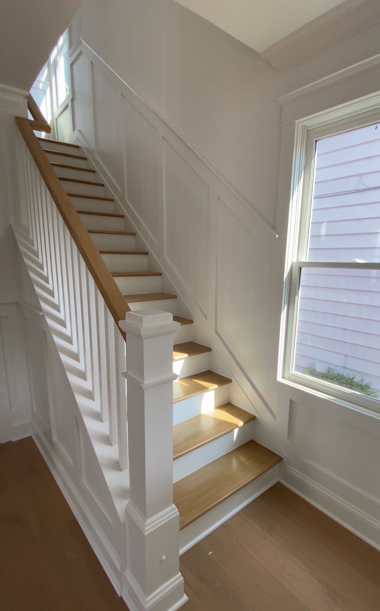 Staircase with light wood steps, white railing and paneling, and a window letting in sunlight.