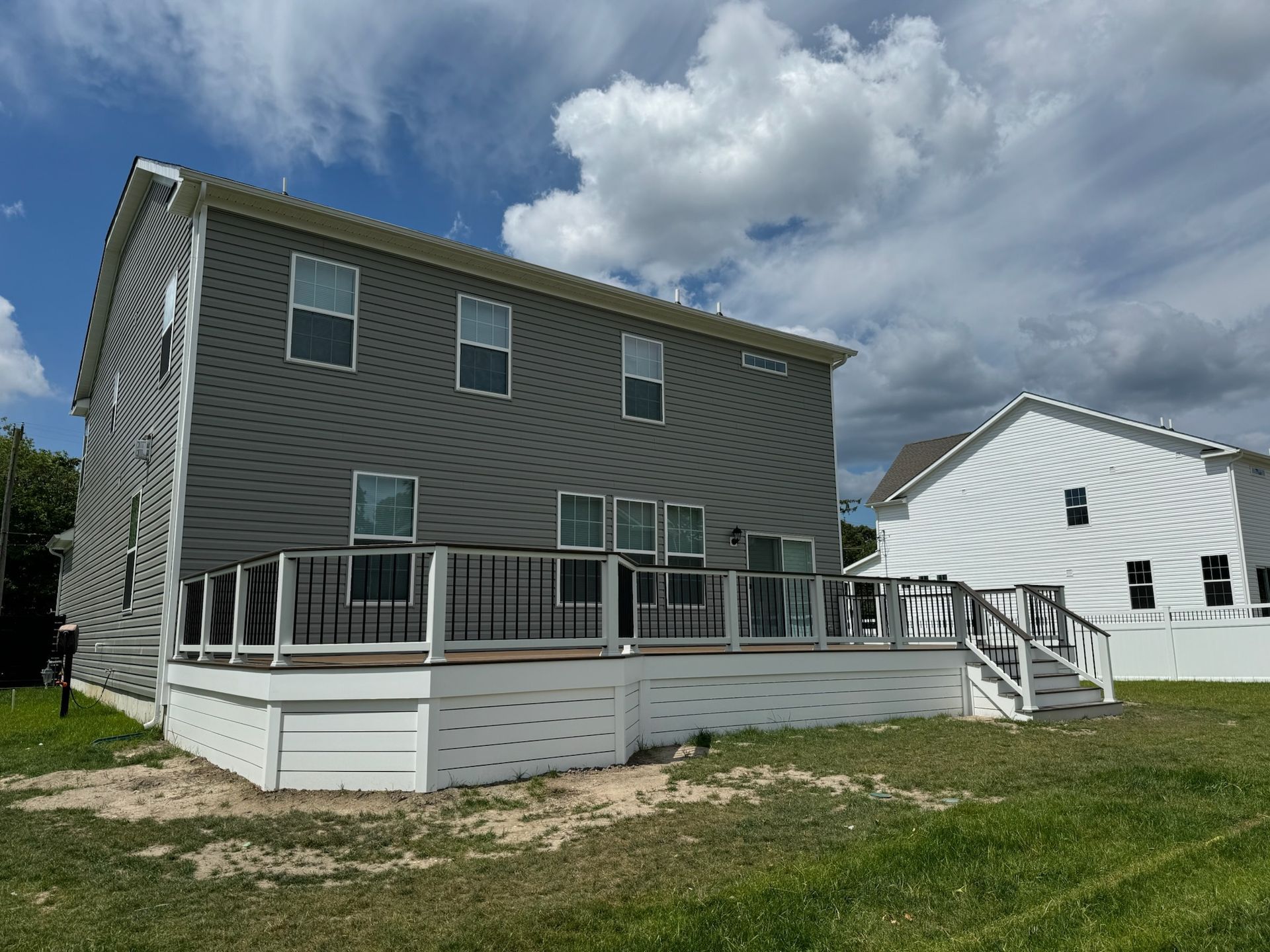 Two-story gray house with a white deck, railings, and steps. Another white house is visible. Sunny day with clouds.