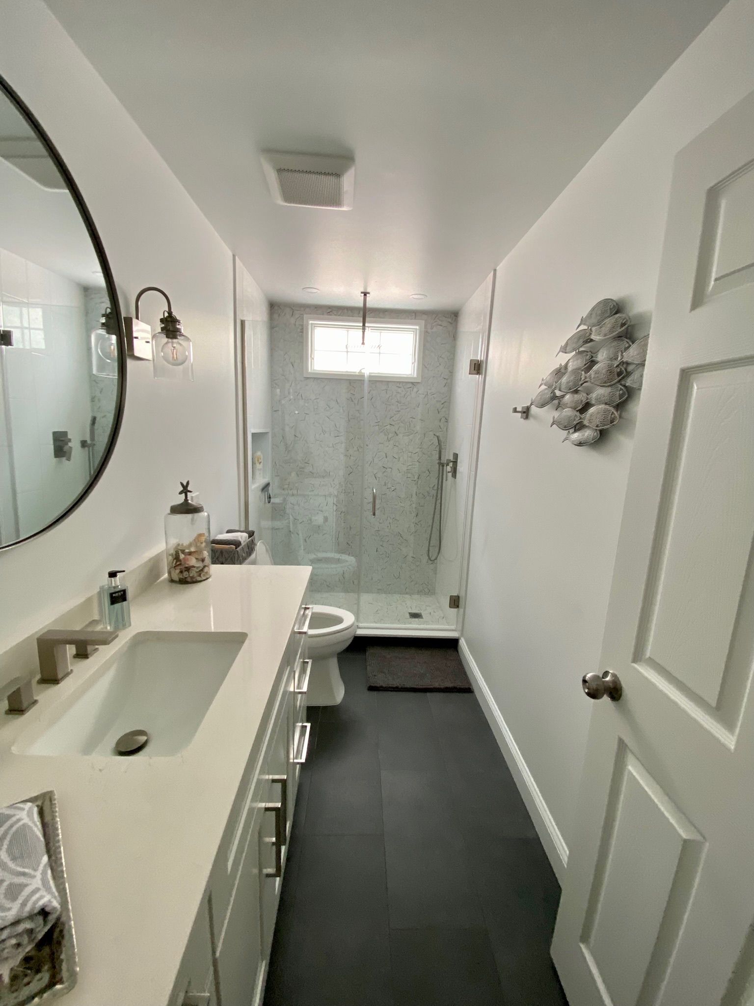 Narrow white bathroom with black floor, vanity with large mirror, and glass-enclosed shower.