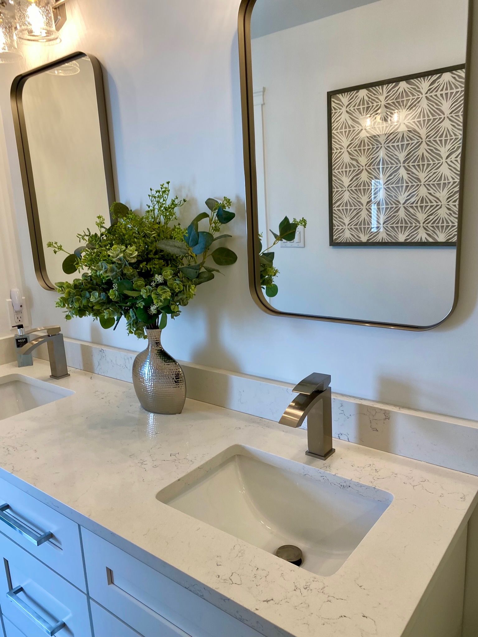 Bathroom with white countertop, two sinks, silver faucets, mirrors, and a floral arrangement.
