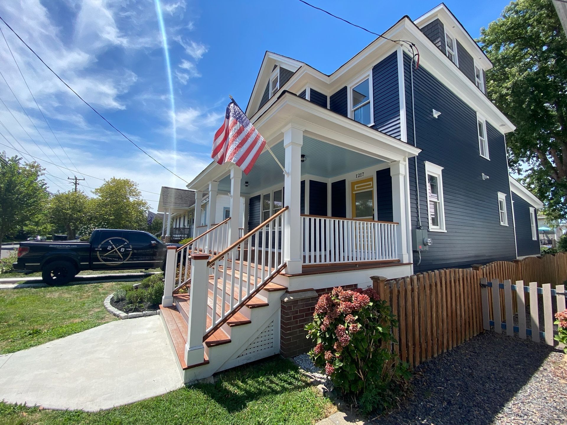 Blue house with white trim, porch, American flag, wooden fence, and black truck.