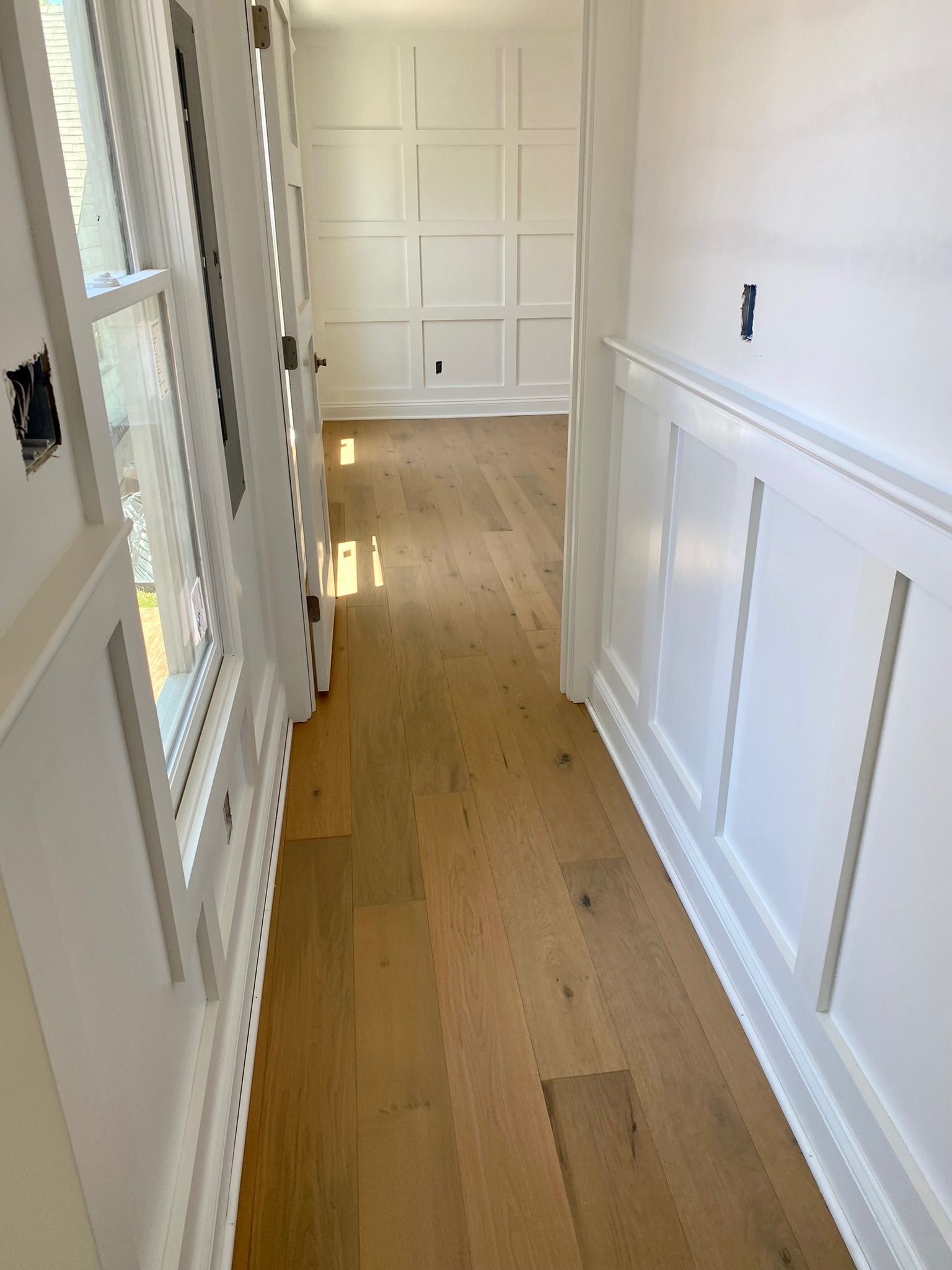 Hallway with light wood floors, white paneled walls, and a door on the left.