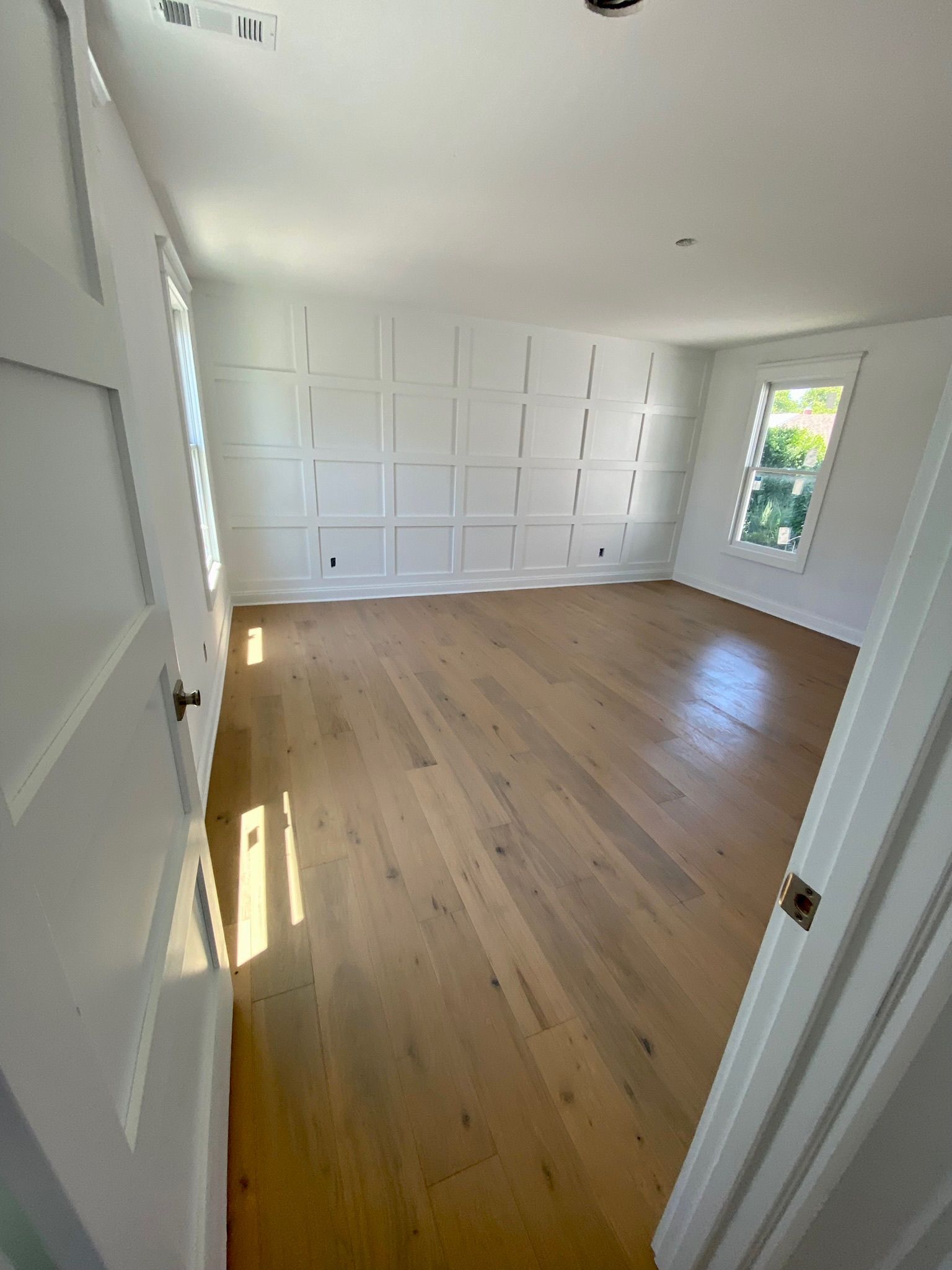 Empty white-walled room with hardwood floors. Paneled feature wall, two windows, and a doorway.