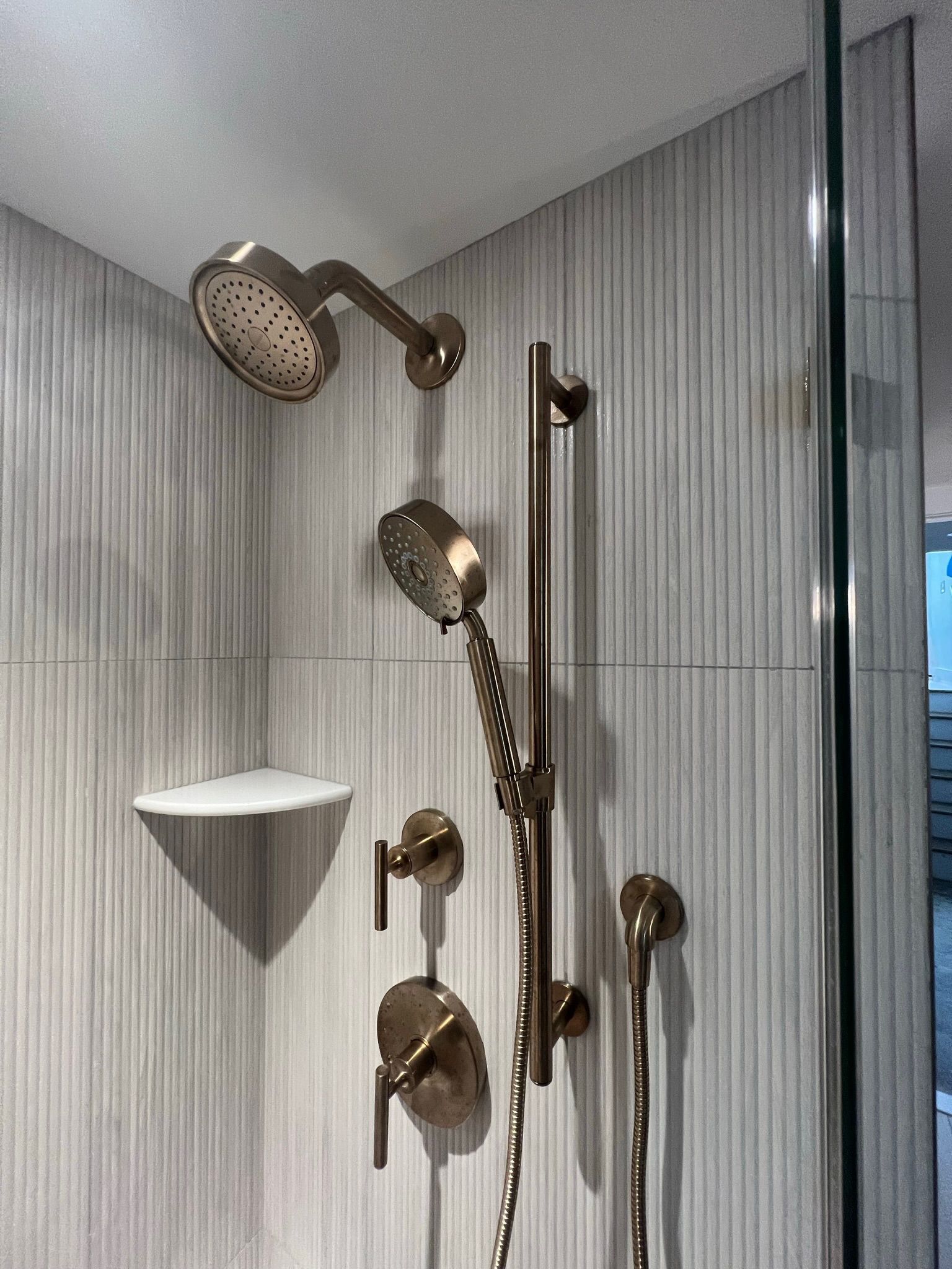 Shower with bronze fixtures against a textured, light gray tiled wall; includes a shelf and glass enclosure.