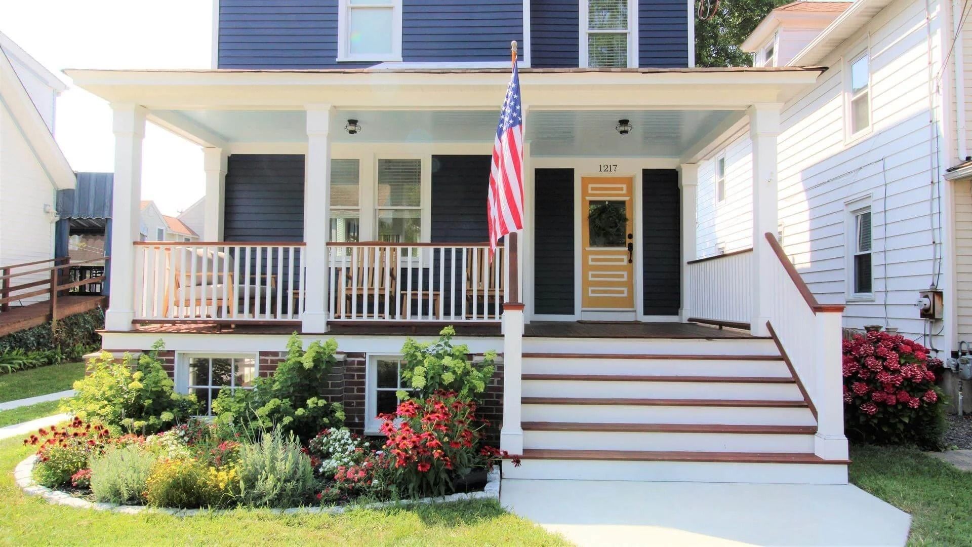 Blue house with porch, American flag, flower garden, white railings, and steps.