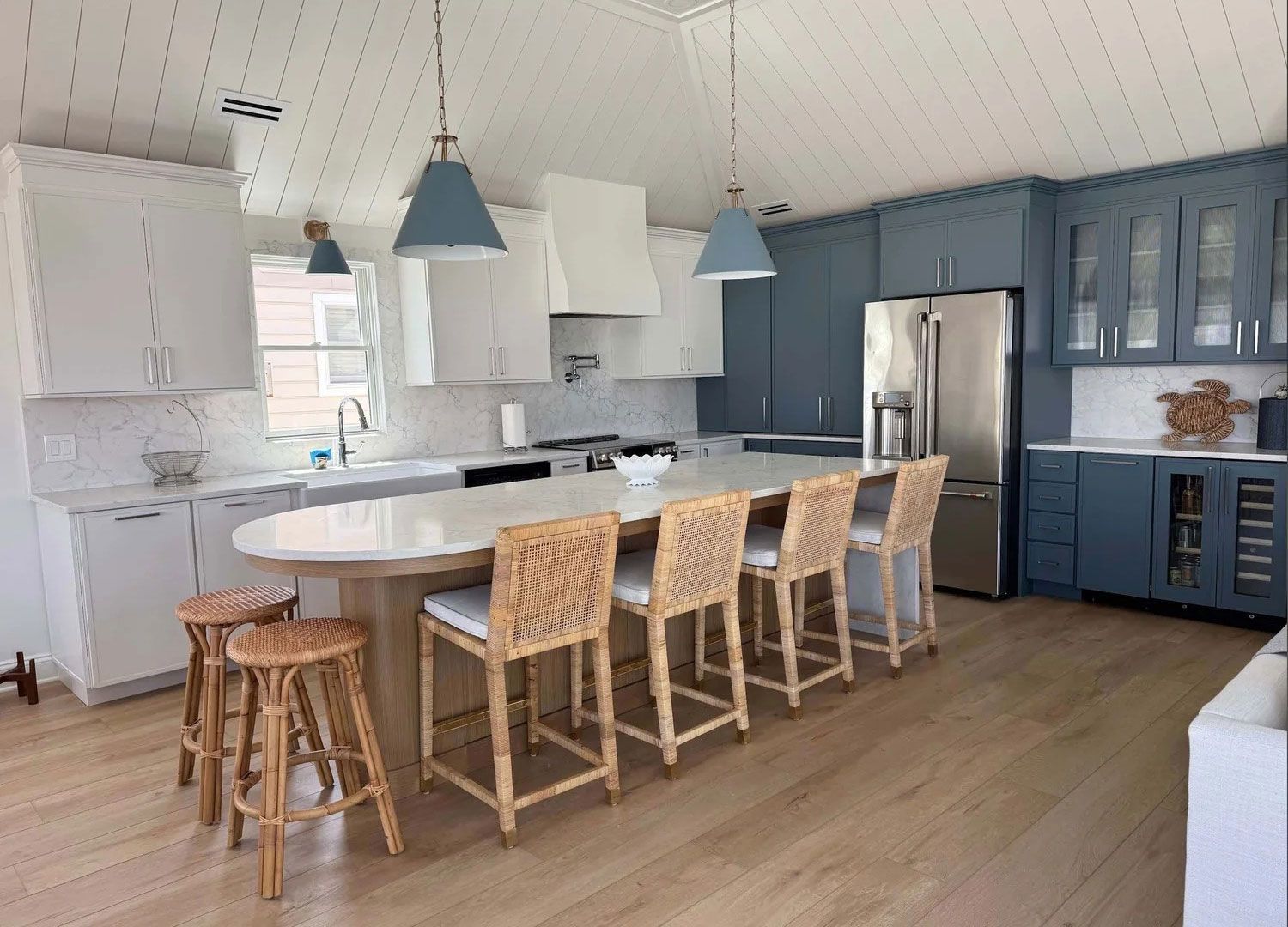Coastal kitchen with blue and white cabinets, large island with rattan stools, and light wood floors.