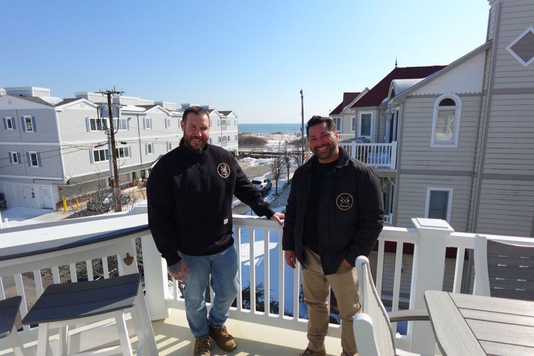 Two men in dark jackets stand on a balcony overlooking a snowy street and the ocean on a bright, sunny day.