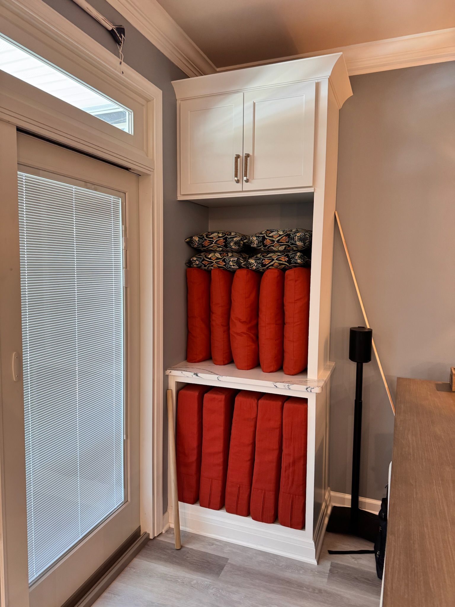 A white cabinet with open shelves holds stacked red seat cushions and patterned pillows, beside a glass-paned door.