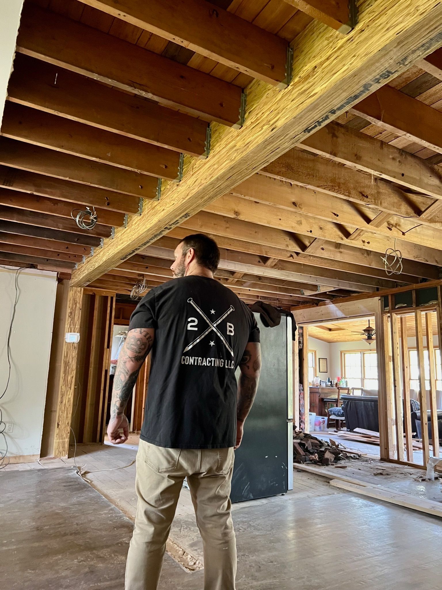 A person with tattoos on their arms stands in a room undergoing construction, looking toward exposed wooden ceiling beams.