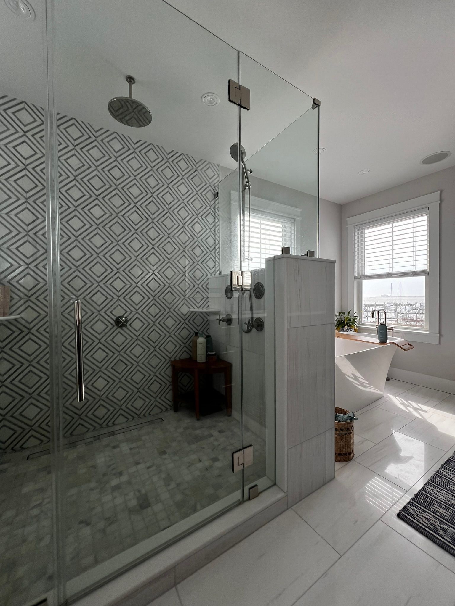 A modern bathroom featuring a glass-enclosed shower with geometric-patterned tile next to a white freestanding bathtub.