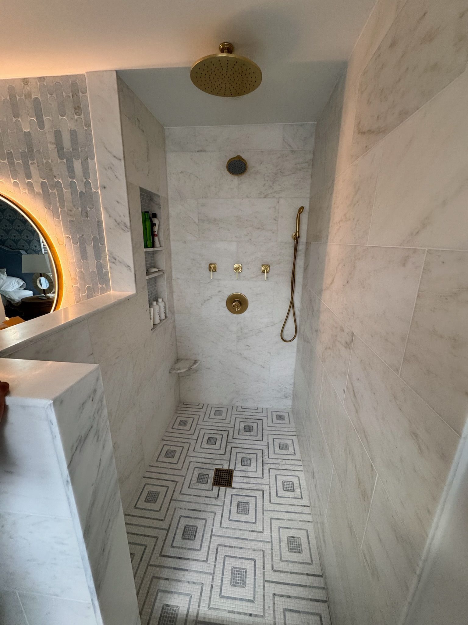 A modern walk-in shower with light marble walls, a geometric tile floor, gold hardware, and an integrated wall niche.