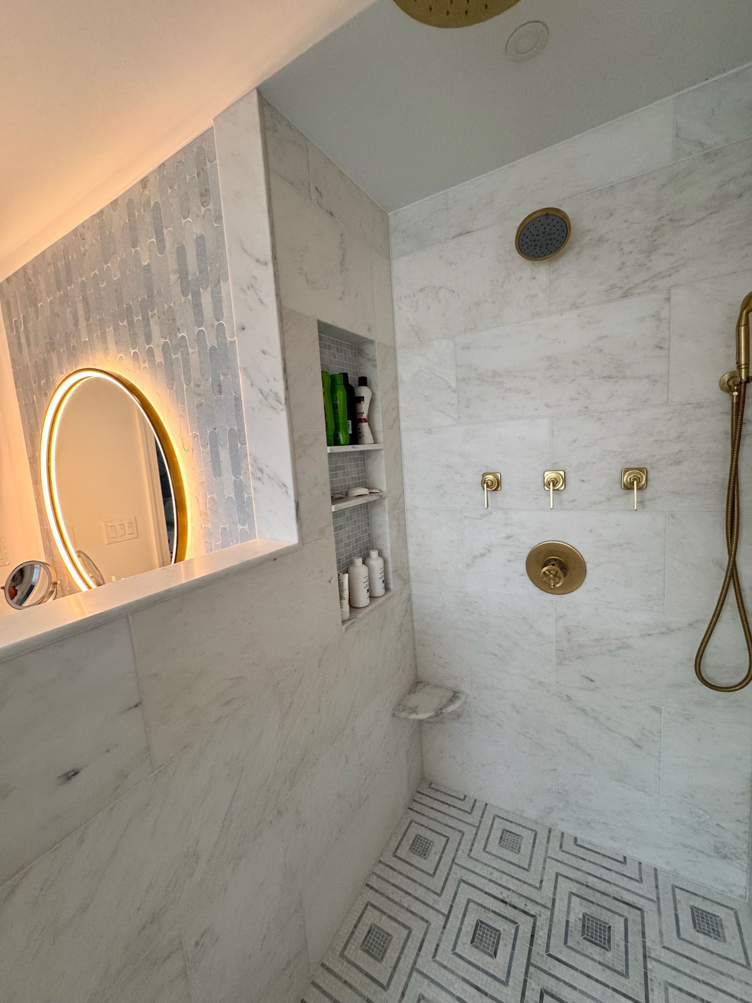 A modern shower with white marble walls, gold fixtures, a built-in niche, and patterned geometric floor tiles.