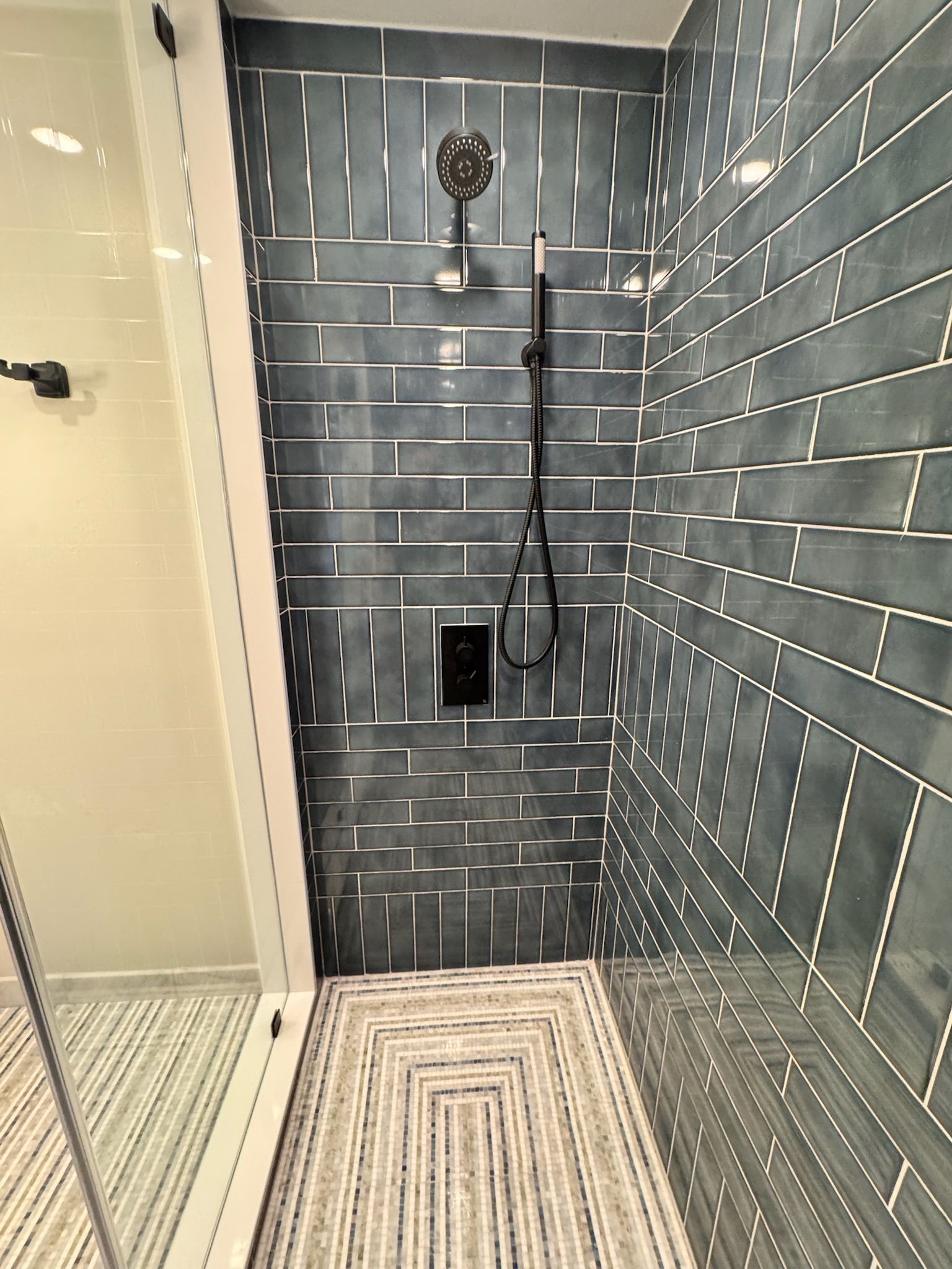 A shower stall with dark blue subway tiles, a black rainfall showerhead, and a hand shower on a patterned tile floor.