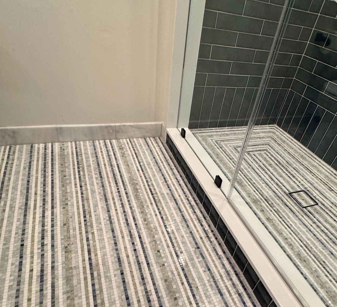 A bathroom floor with grey-striped carpeting meeting a white marble threshold and a glass-enclosed shower with tiled floor.