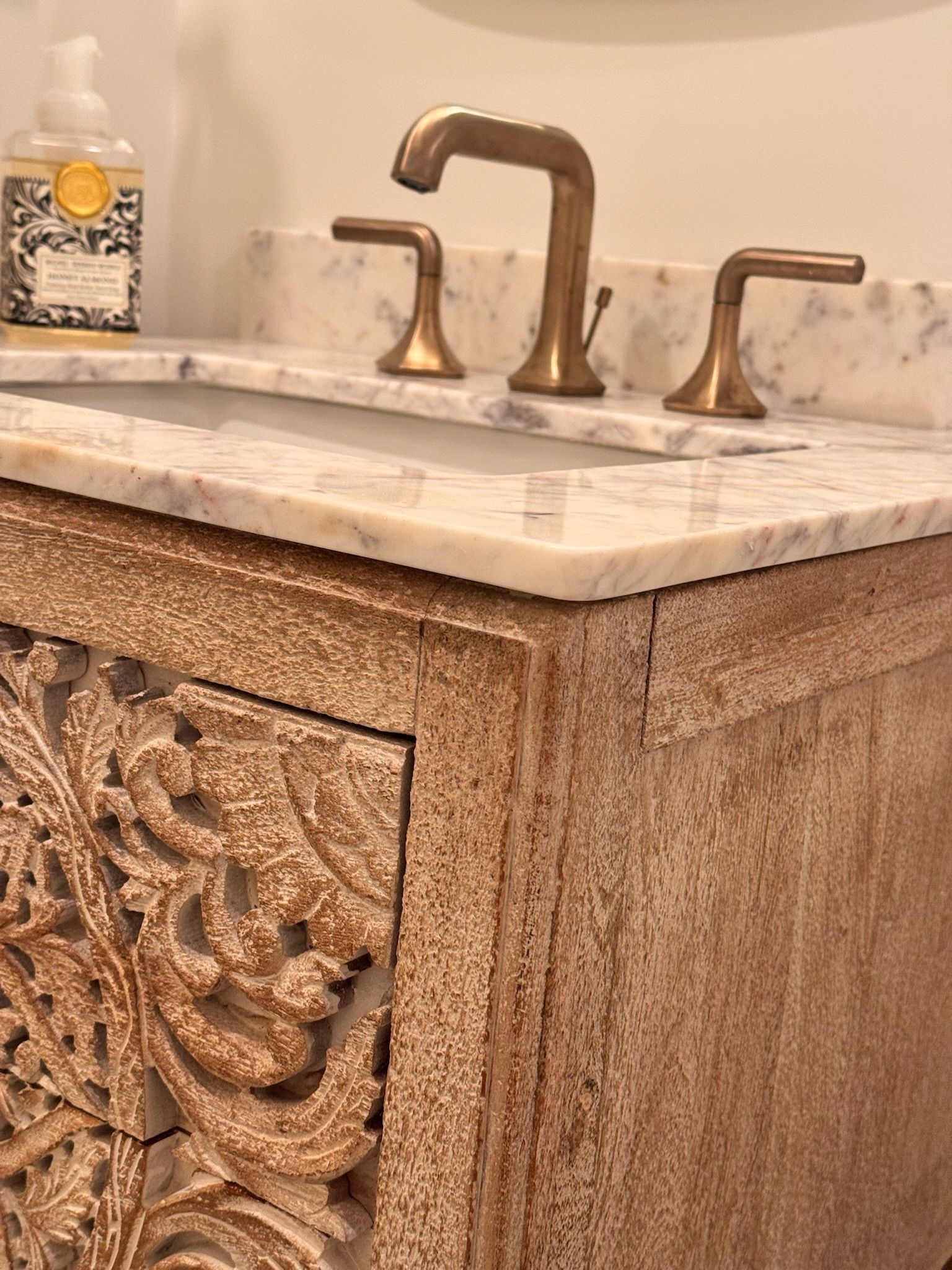 Close-up of a rustic carved wooden bathroom vanity with a marble countertop, brass faucet, and a soap dispenser.
