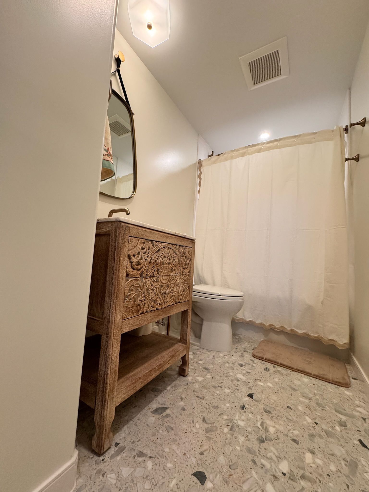 A bathroom with a decorative carved wood vanity, terrazzo floor, a mirror, and a neutral-colored shower curtain.