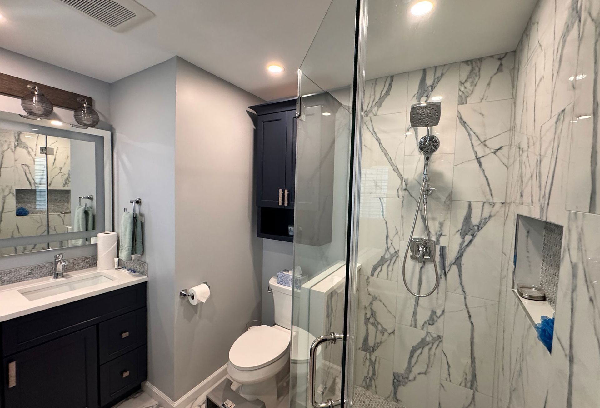 Modern bathroom with a white marble walk-in shower, dark blue vanity, gray walls, and a toilet under a wall cabinet.