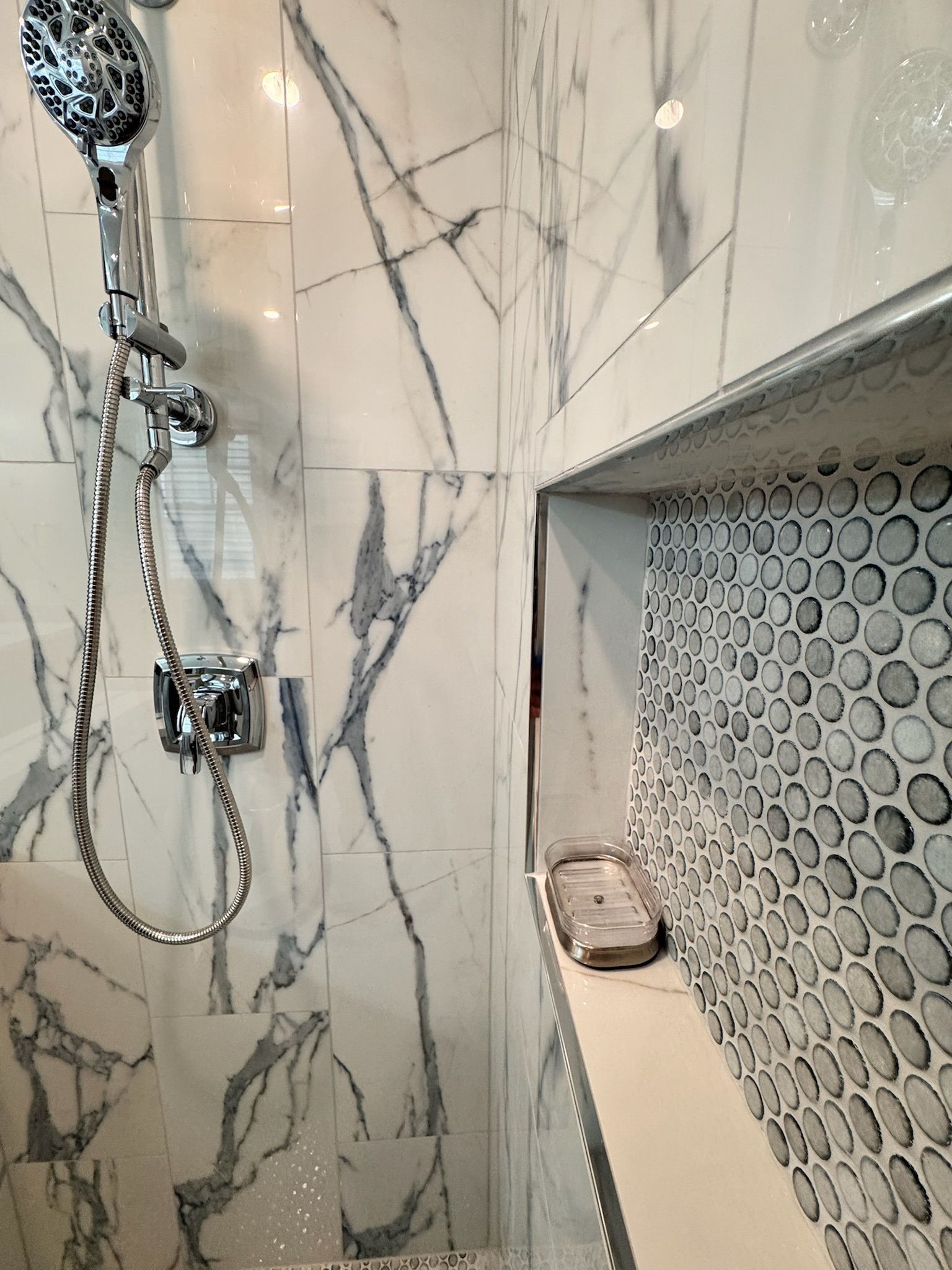 Shower wall with white marble-look tiles, a chrome hand-held shower head, and a recessed niche featuring pebble tile.