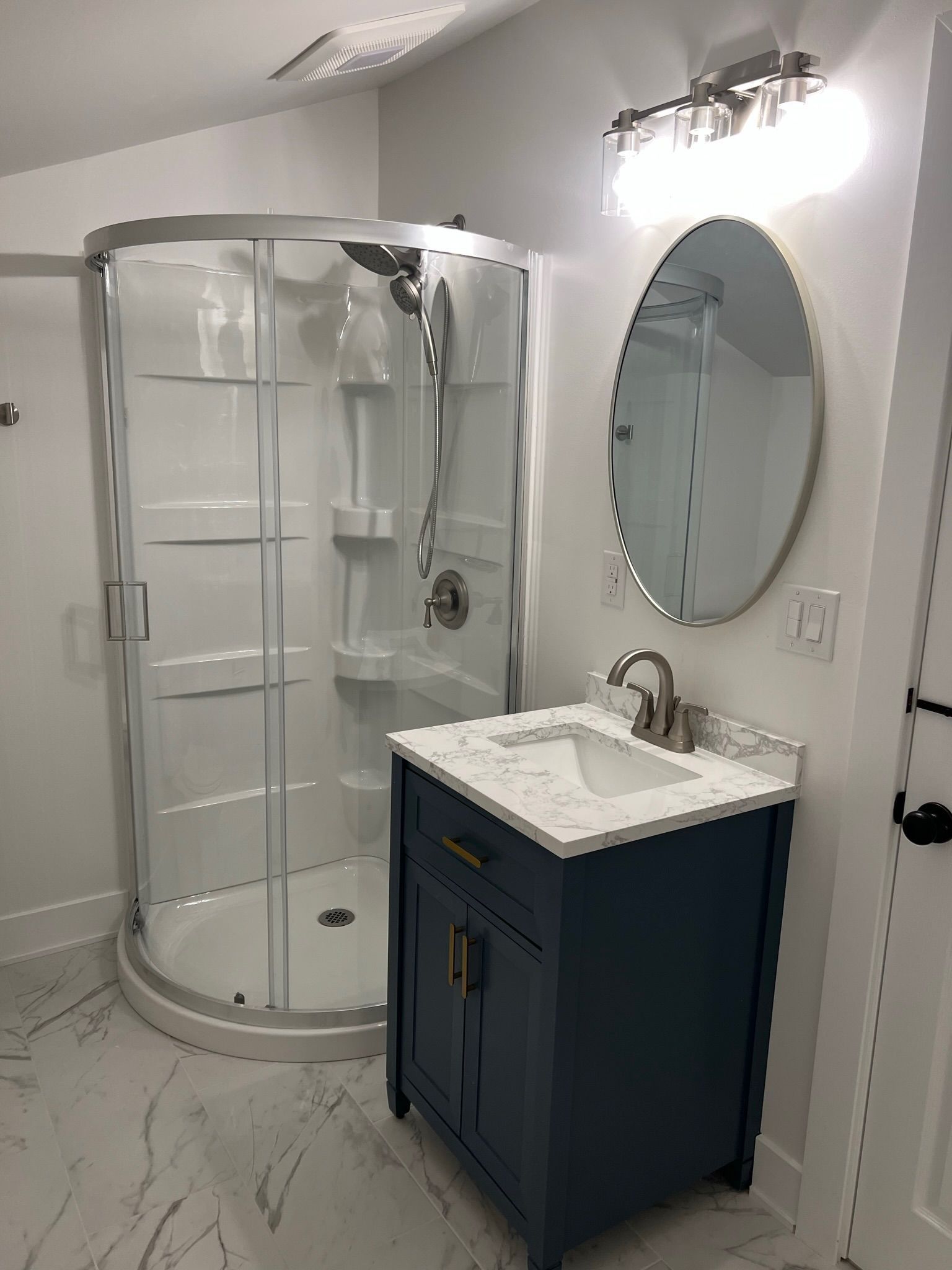 A small bathroom features a navy blue vanity with a white marble top, a corner glass shower, and an oval mirror.