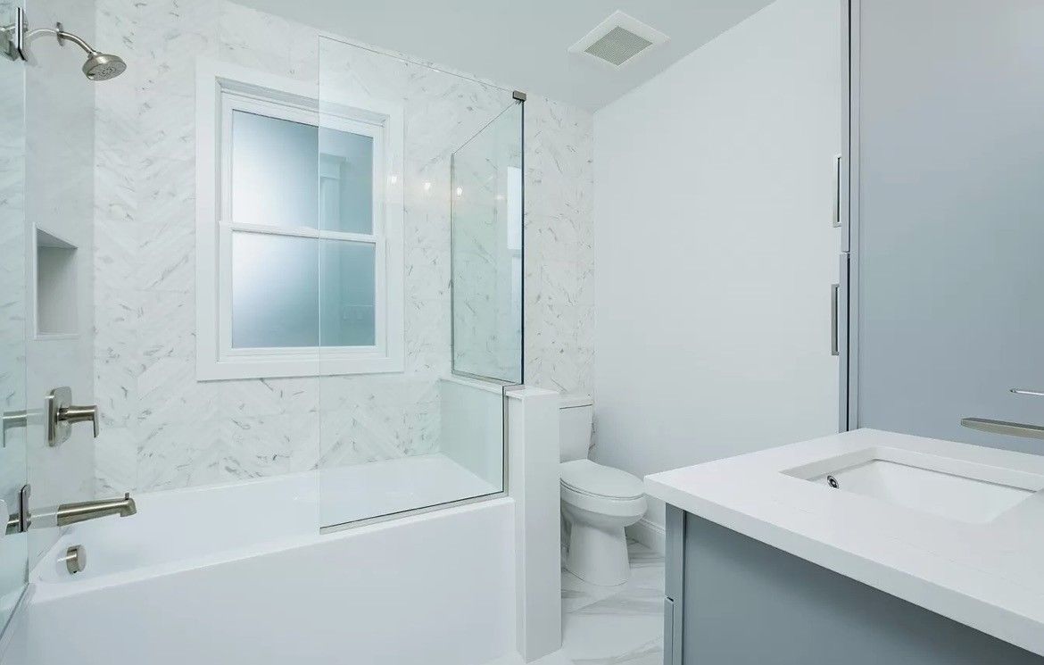 A white, modern bathroom with a marble-tiled bathtub, glass shower screen, toilet, and vanity sink.