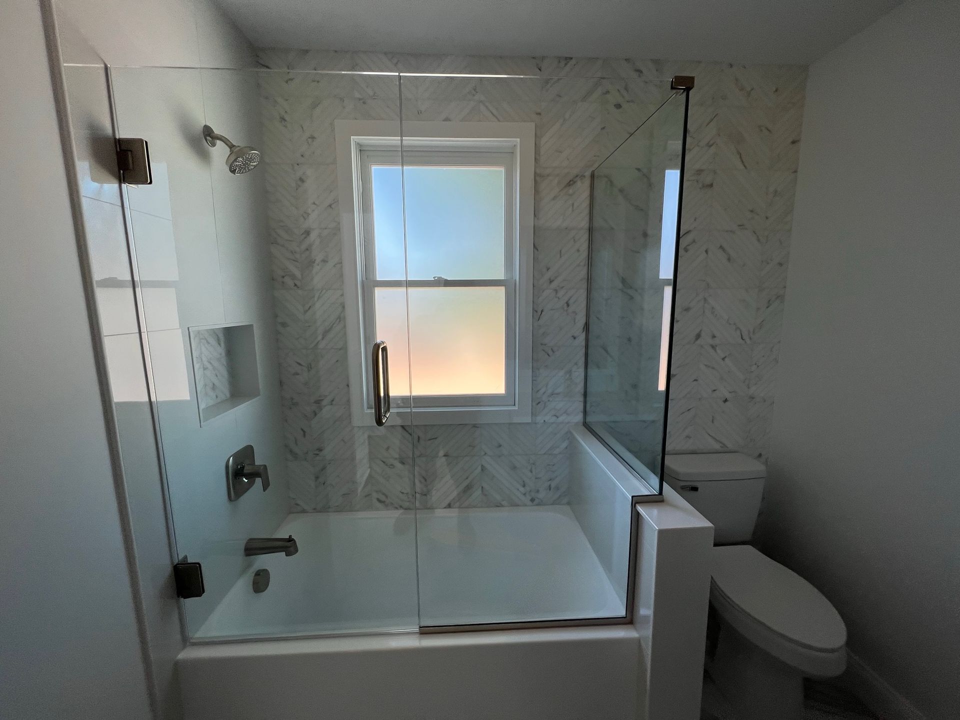 A bathroom with a white bathtub, marble-patterned tile walls, a glass shower enclosure, and a toilet to the right.
