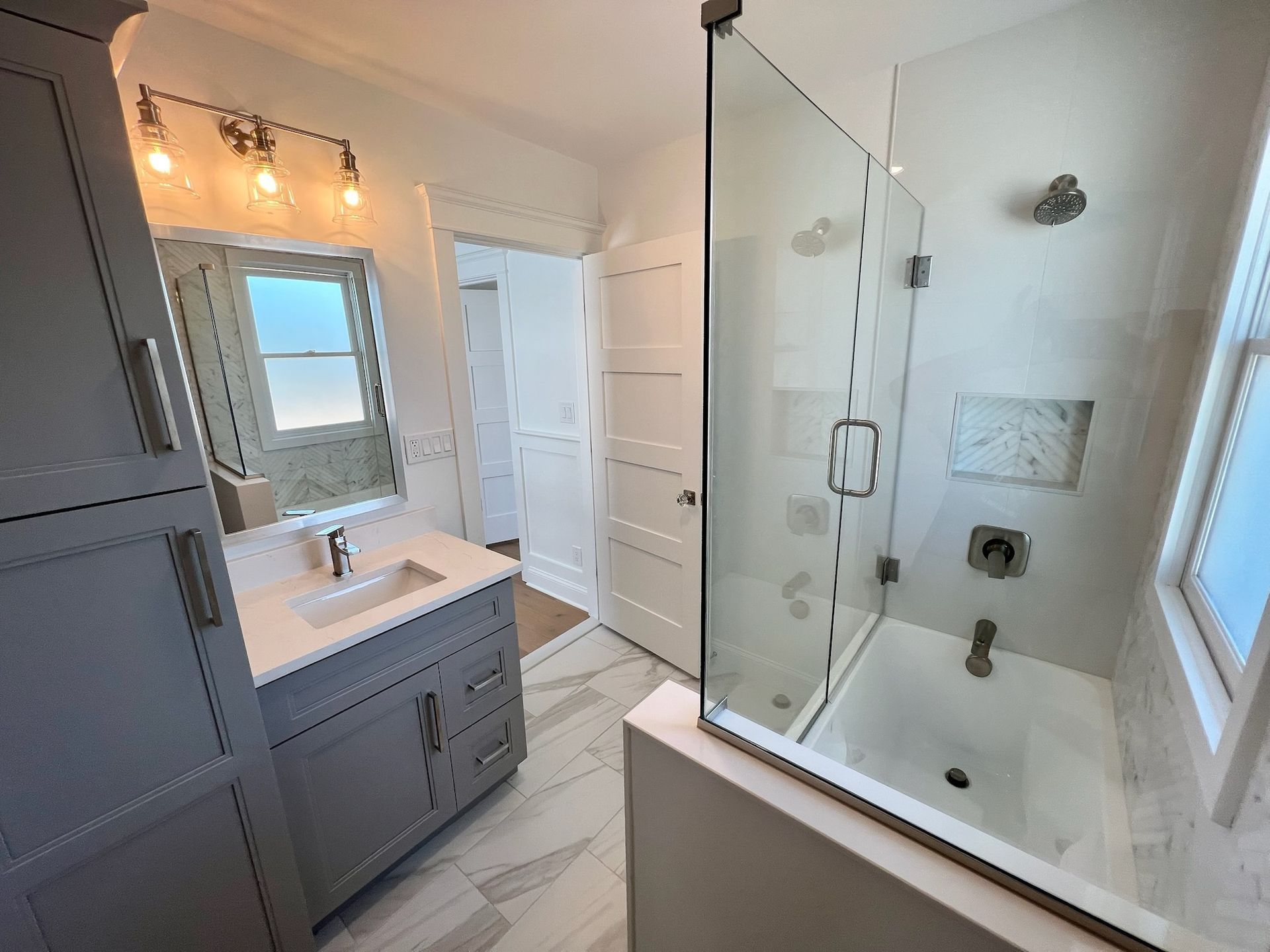 A modern, bright bathroom with a gray vanity, marble tile floor, and a glass-enclosed shower-tub combination.