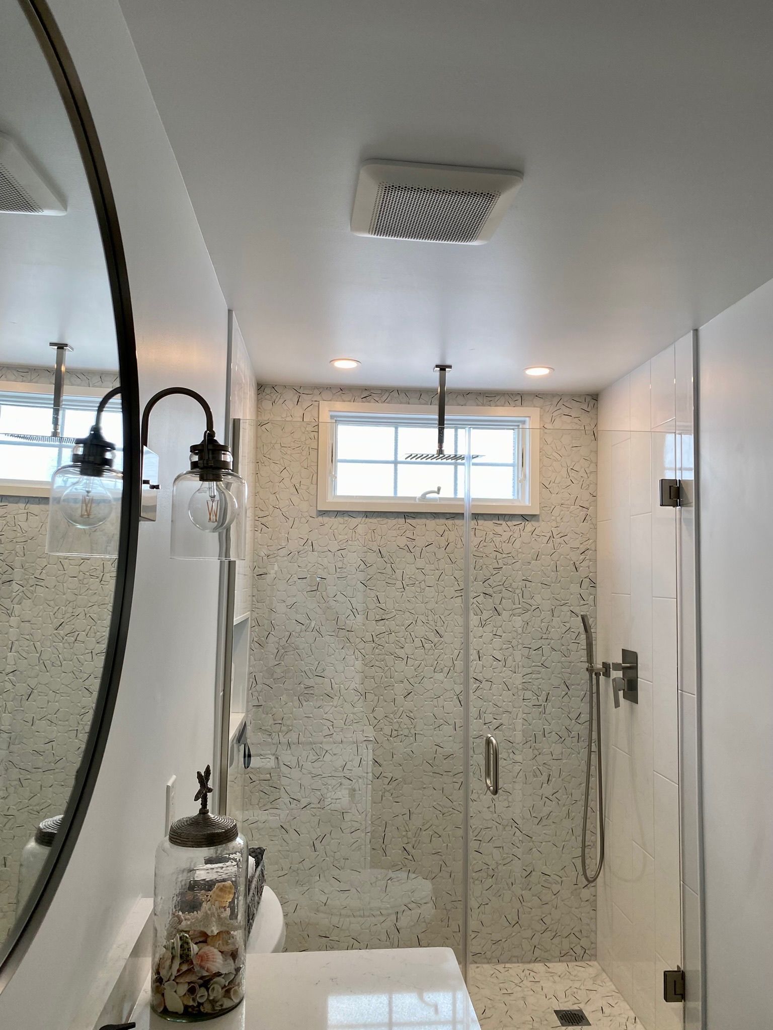 A bathroom vanity with a mirror next to a walk-in shower featuring white walls and a speckled tile floor and backsplash.
