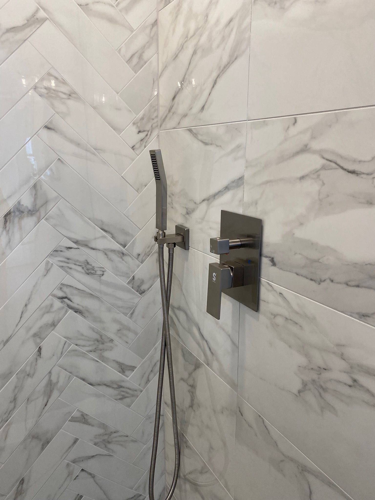 Modern chrome shower faucet with a handheld wand installed against a white and grey marble herringbone tiled wall.