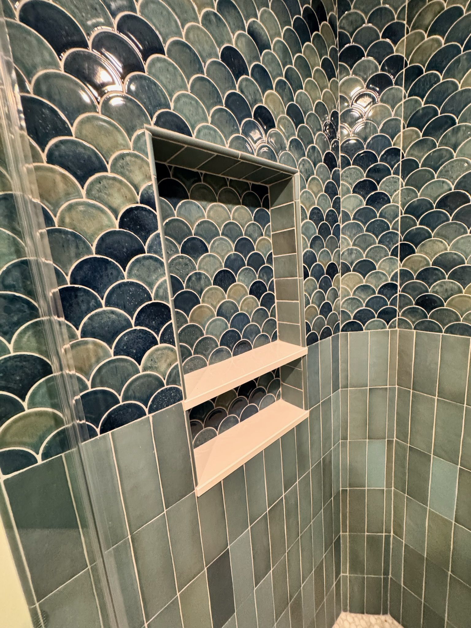 A shower wall features a vertical transition from dark green rectangular tiles to blue and green scalloped scale tiles.