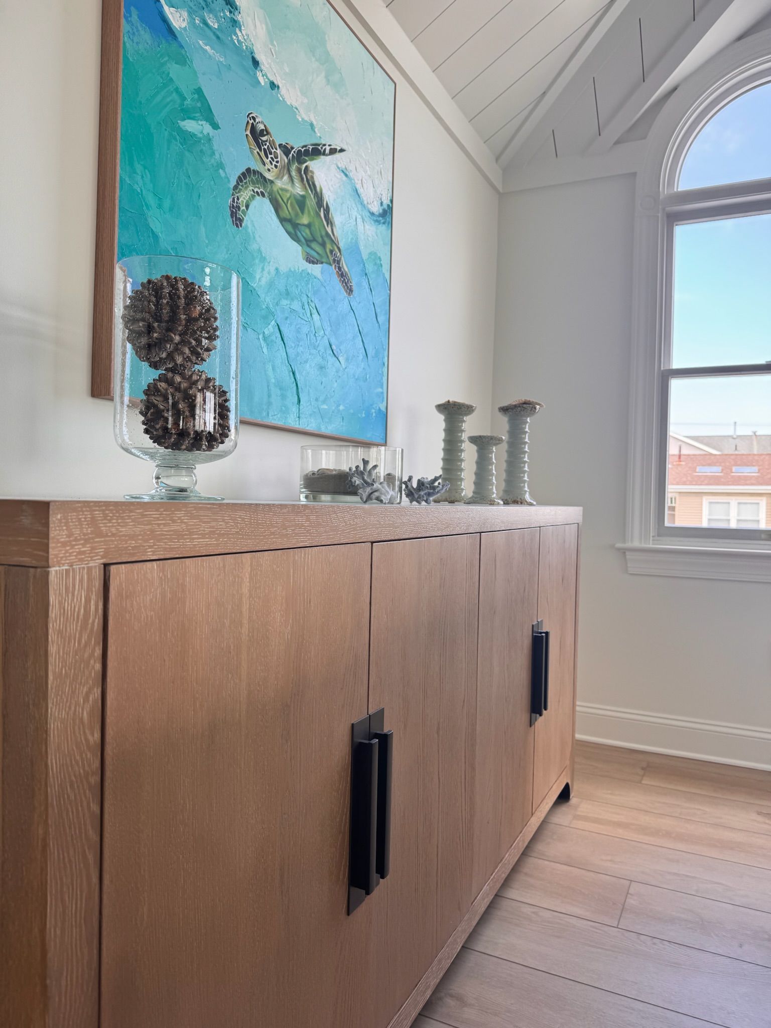 A wood cabinet with black handles stands beneath a painting of a sea turtle, with decorative items on the surface.