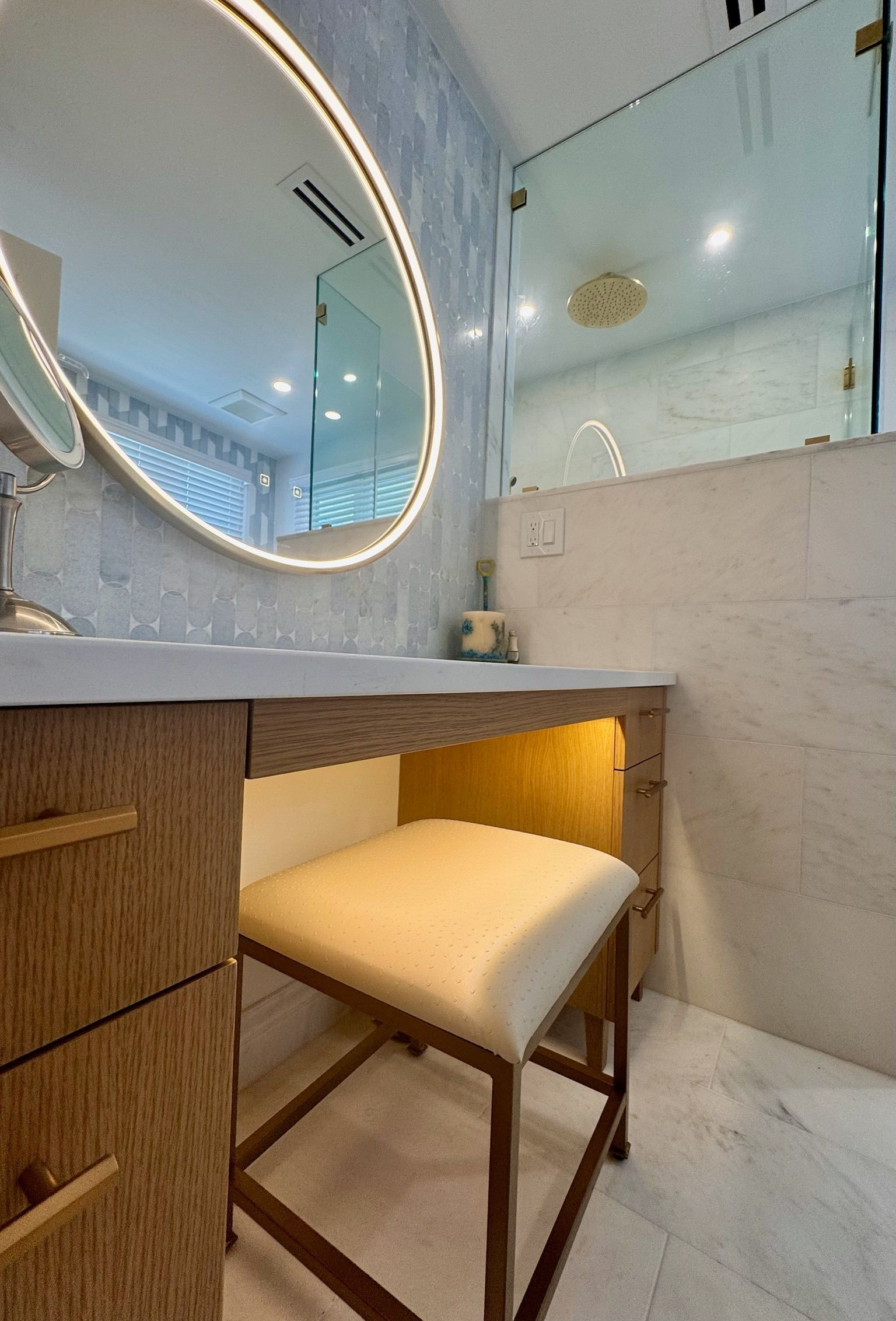 A modern vanity with a wood-toned cabinet, an oval LED-lit mirror, and a cushioned stool in a marble-tiled bathroom.
