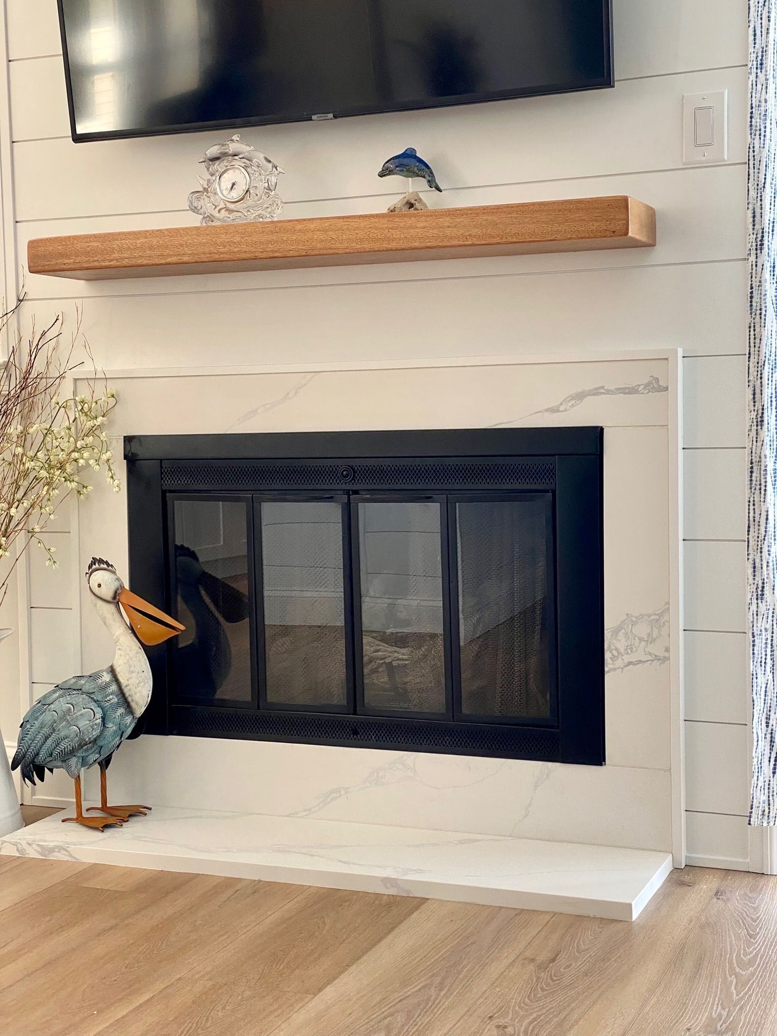 A fireplace with white marble tiling and a black iron screen, topped by a wooden mantel shelf and a wall-mounted TV.