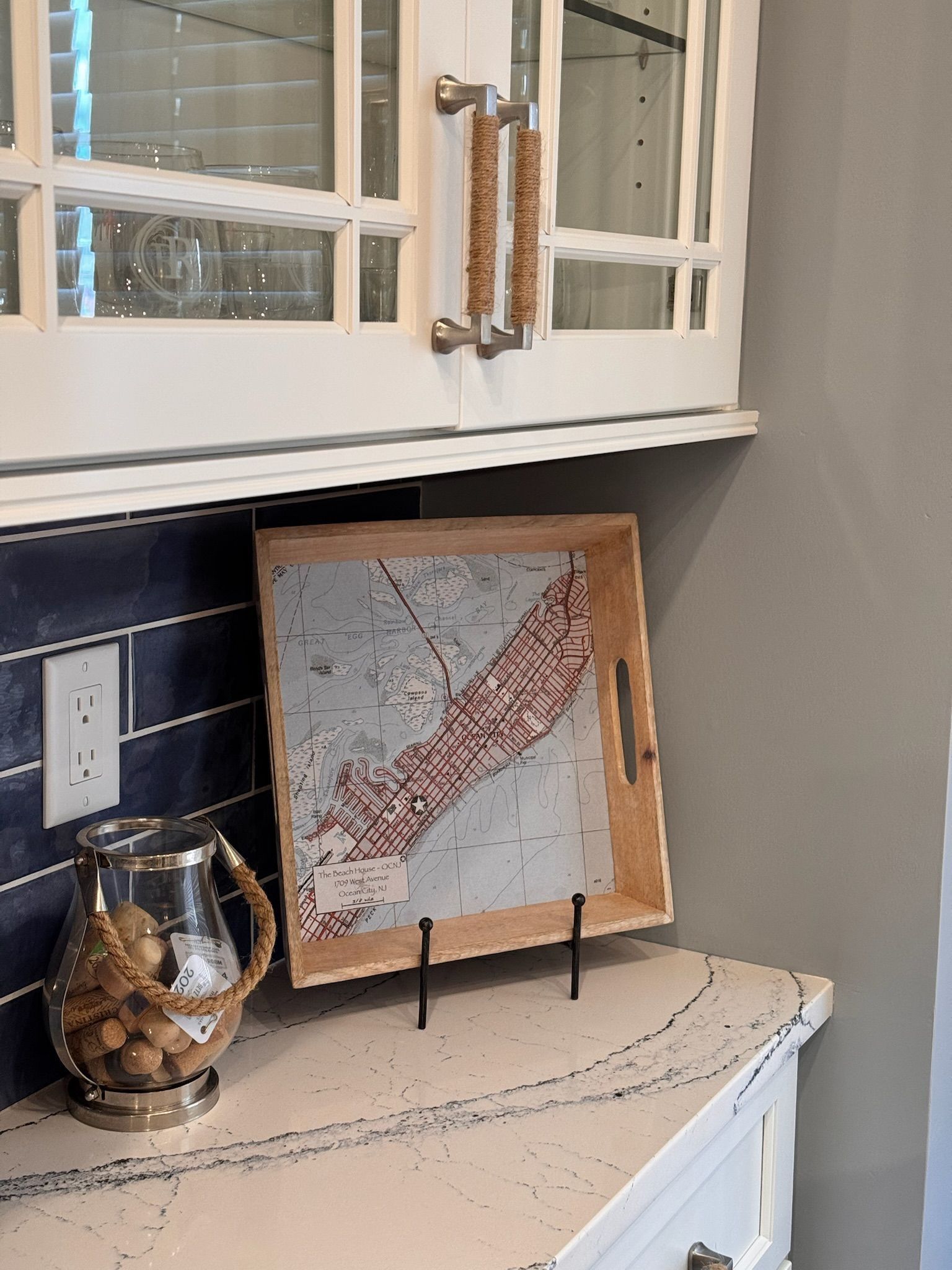 A wooden tray with a map print rests on a speckled countertop next to a glass jar filled with small objects under cabinets.